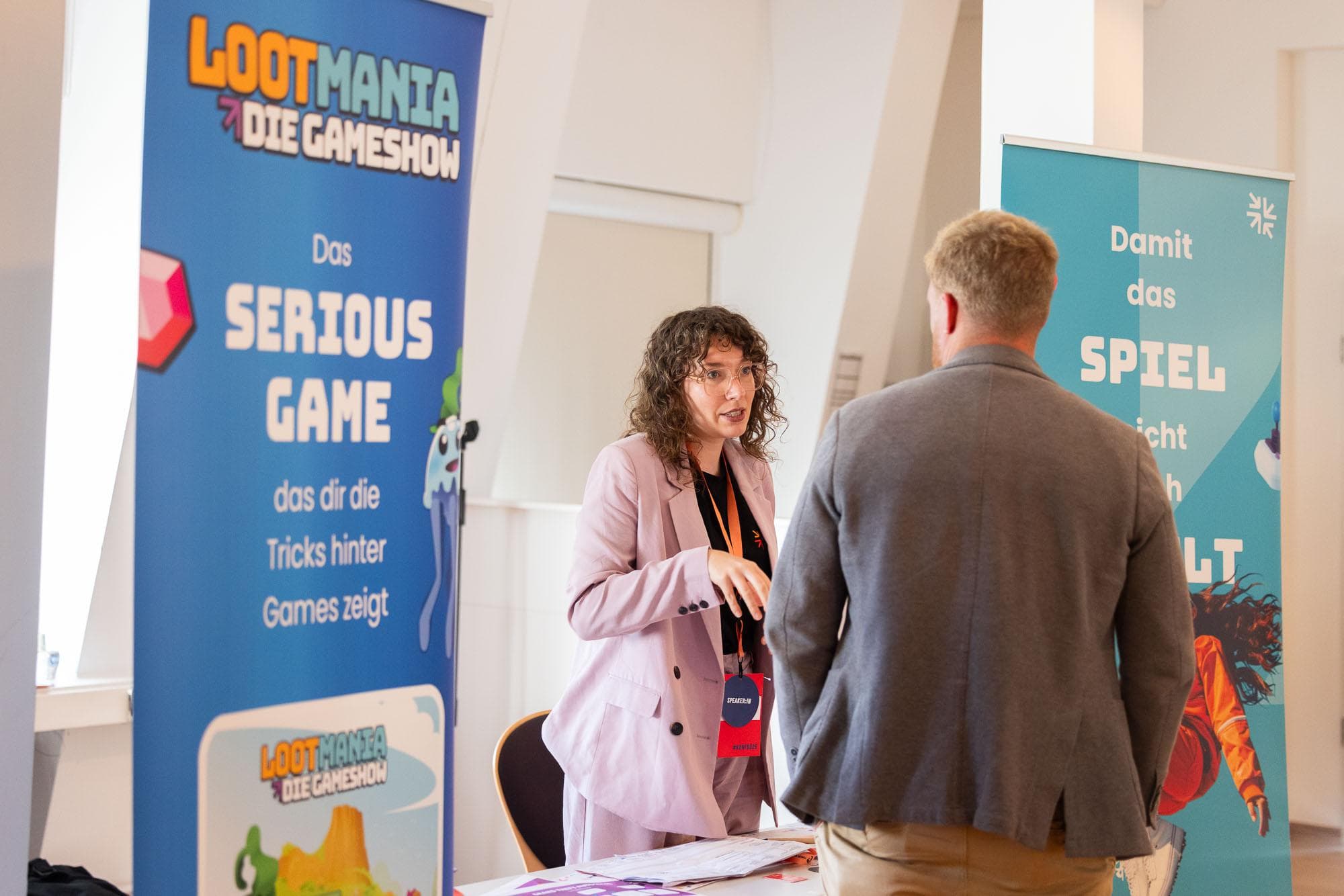 Eine GAMESHIFT-Mitarbeiterin spricht an einem Messestand zwischen zwei Projektbannern mit einem Besucher.
